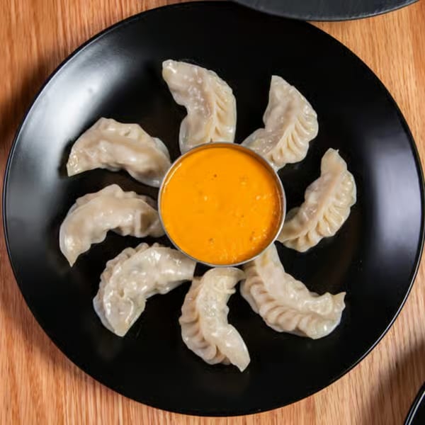 A black plate of Himalayan Wild Yak momos arranged around a small bowl of orange dipping sauce.