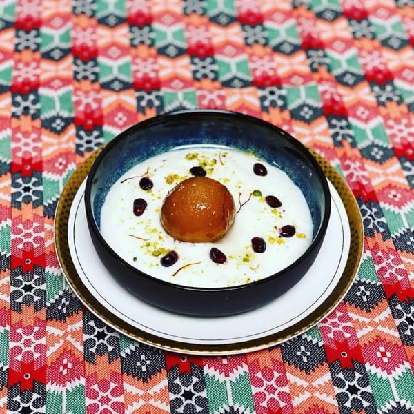 Gulab Jamun dessert in a dark bowl with white cream, garnished with pistachios and saffron threads.