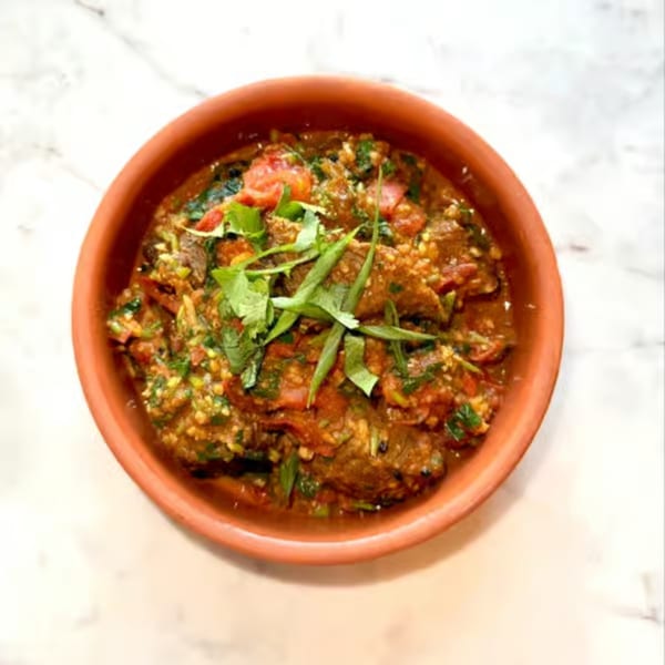 Nepalese curry with meat, tomatoes, and green herbs in a rustic terracotta bowl on a marble surface.