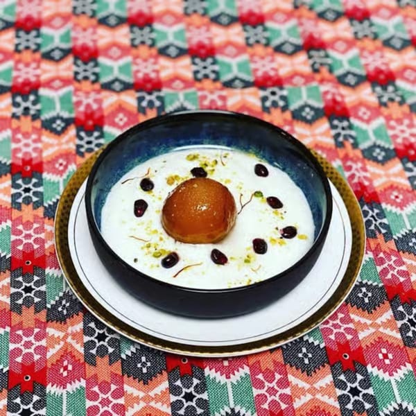 Gulab Jamun dessert in a dark bowl with cream, saffron, and dried fruit on a colorful tablecloth.